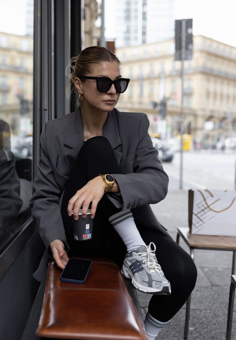Grå oversize blazer, svarta leggings, vita strumpor med gröna ränder, grå sneakers, svarta solglasögon, guldklocka, kaffekopp, smartphone.