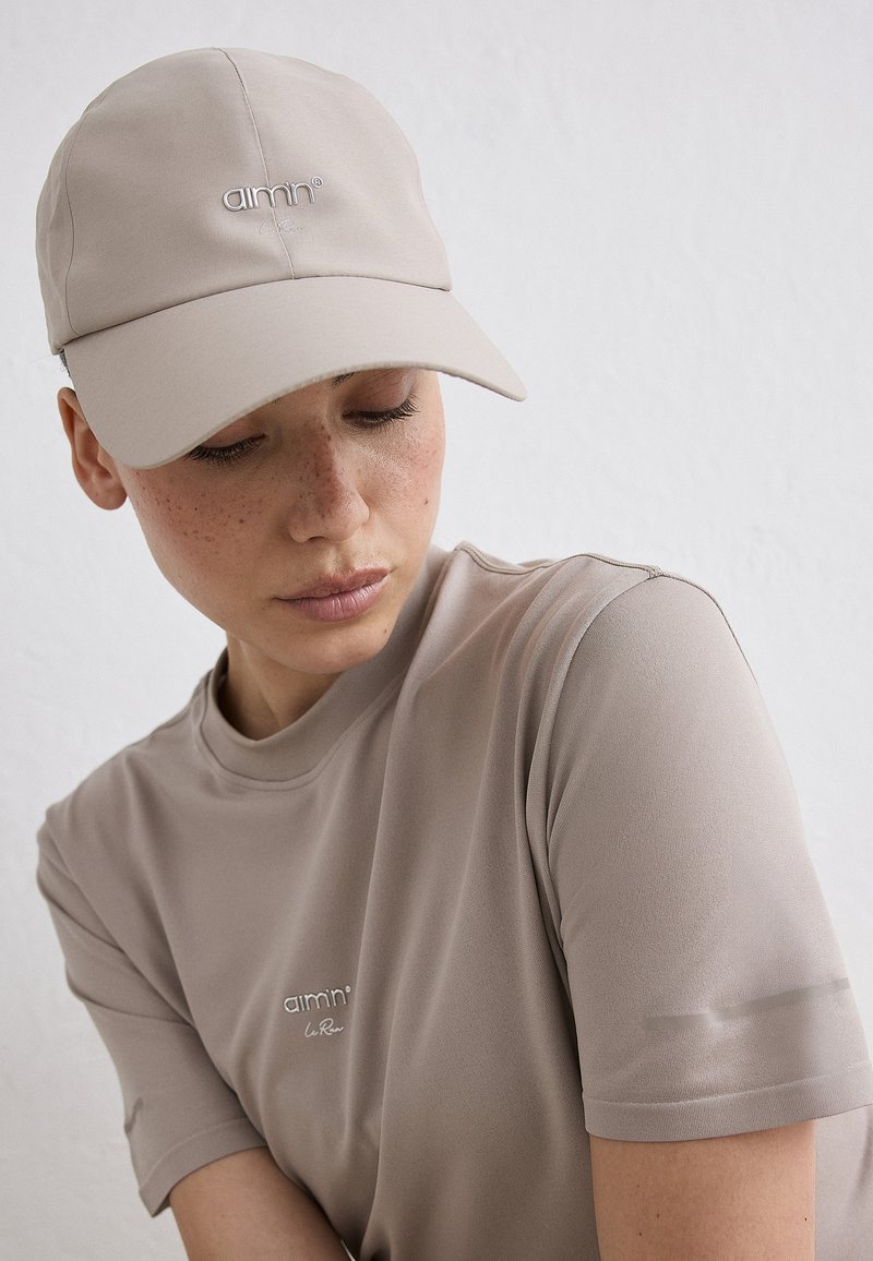 Jeune personne avec des taches de rousseur portant une casquette beige et un t-shirt beige assorti, regardant vers le bas sur un fond clair uni.