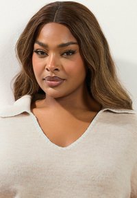 Vaaleanbeige neulepaita, jossa on V-pääntie ja kaulus. Paita on pehmeätekstuurinen, siinä on hienovarainen kuvio ja kontrastivärinen reuna pääntiellä ja helmoissa.