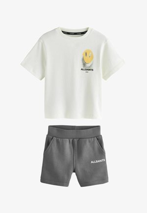 Ung dreng står med hænderne i lommerne iført en hvid t-shirt med smilende ansigt, grå shorts, hvide sokker og hvide sneakers foran en grå baggrund.