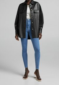 Person i svart imitationläder oversize knäppjacka, vit tanktop, blå skinny jeans och genomskinliga högklackade sandaler.