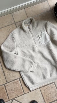 Pulover iz flisa v off-white barvi z zadrgo pri ovratniku, texturirano površino in kontrastno zadrgo na žepu. Rebrasti manšeti in rob.