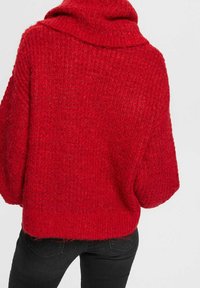 Roter gestrickter Pullover mit hohem, umgeschlagenem Kragen und lockerer Passform. Strukturierter Stoff mit sichtbaren Nähten. Der Saum ist leicht geschwungen.