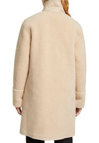 Beige fleecejacka med hög krage, har en mjuk textur och vida ärmar. Bakifrån visas en rak silhuett och rena linjer.