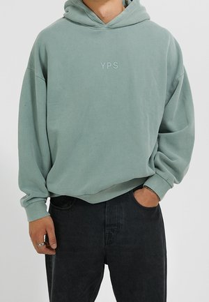 Homme portant un sweat à capuche vert clair avec l'inscription "YPS" sur la poitrine et un jean noir, une main dans la poche et l'autre le long du corps.