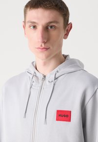 Grå zip-up hettegenser med et rødt rektangulært merke merket "HUGO." Har snøring i hette og myk tekstur. Nærbilde av fronten.