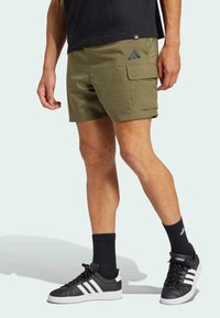 Green cargo shorts with side pocket, worn with black sneakers featuring white stripes and black socks. Made of lightweight fabric.