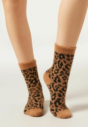 Chaussettes brunes en peluche avec un motif léopard noir. Le haut présente un revers côtelé doux. Le matériau semble moelleux et texturé.