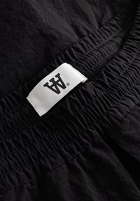 Ceinture élastique noire avec texture froncée, munie d'une étiquette blanche affichant le logo de la marque. Le matériau semble léger et lisse.