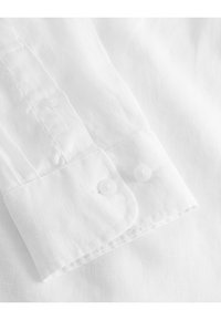 Polsino di camicia in lino bianco con due bottoni trasparenti, texture liscia e dettagli di cucitura discreti lungo il bordo.
