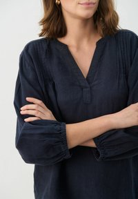 Tamno plava lanena bluza s v-izrezom, teksturiranim ramenima i elastičnim napuhanim rukavima, koja prikazuje opušteni, lagani dizajn.