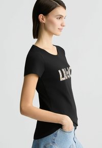 Juoda trumpa rankovė t-shirt, dekoruota blizgučiais "LUJO" raštu priekyje. Lygus tekstūra, apvalus kaklo iškirpimas ir prigludusi siluetas.