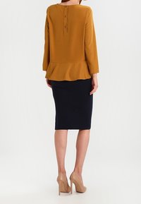 Top peplum senape con chiusura a bottoni sul retro, abbinato a una gonna a matita blu navy e decolleté nudi, che mette in mostra una texture di tessuto liscia.