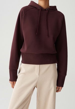 Osoba ubrana w ciemnoczerwony kaptur z troczkami oraz kremowe, szerokie spodnie z prążkowanej tkaniny, stojąca na jednolitym tle.