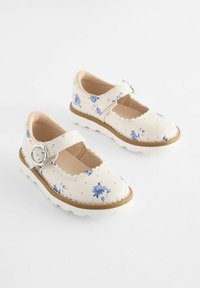 Weiße Mary Jane Schuhe mit Blumenmuster, gewelltem Rand, verstellbarem Riemen und einer Gummisohle mit braunen Akzenten.
