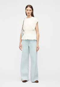 Mujer con top blanco sin mangas con peplum, vaqueros de pierna ancha azul claro con adornos, zapatos de estampado de leopardo y bolso plateado de hombro.