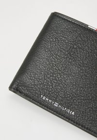 Zwarte leren portemonnee met een gestructureerde afwerking. Kenmerkt zich door zilveren branding "TOMMY HILFIGER" gedrukt in de rechterbenedenhoek. Vouwmodel.