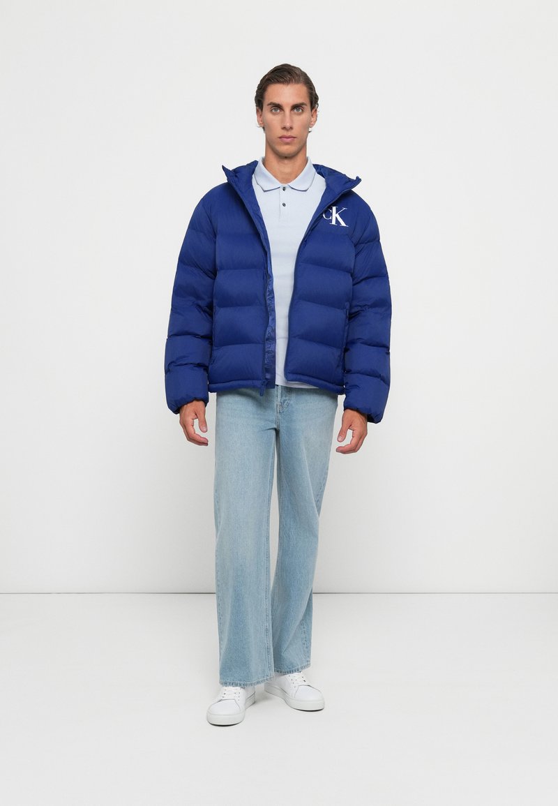 Egy férfi áll, kék puffertakart viselve, világoskék pólóval, laza világoskék farmernadrággal és fehér tornacipővel egy sima fehér háttér előtt.