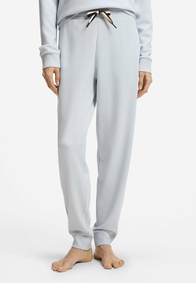 Personne portant un pantalon de jogging gris clair avec un cordon noir et beige, debout pieds nus devant un fond blanc uni.