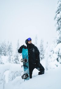 Giacca da snowboard nera, snowboard blu con attacchi neri e mimetici, sfondo innevato, alberi in lontananza, cielo coperto.