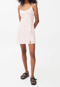 Frau trägt ein weißes Minikleid mit roten Punkten, dünnen Trägern, einem leichten Schlitz vorne und schwarzen, gesteppten Sandalen mit Schleifendetails.