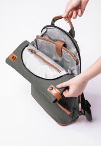 Grauer Rucksack mit einem hellbraunen Ledergriff und Akzenten. Offen, um ein kariertes Innenleben zu zeigen. Verfügt über einen Reißverschluss und einen äußeren Druckknopf.