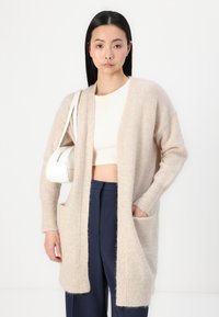 Cardigan beige realizzato in morbido materiale peloso, con apertura frontale, maniche lunghe, due tasche frontali e una vestibilità comoda.