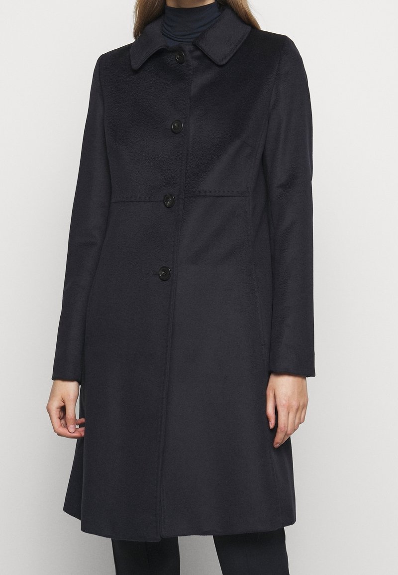 Femme portant un long manteau noir boutonné avec un col par-dessus un col roulé noir, debout devant un fond clair uni.