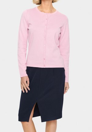 Kobieta ubrana w różowy rozpinany sweter i granatową spódnicę do kolan z przednim rozcięciem, stojąca na jednolitym tle.