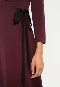 Bordowa sukienka z teksturowanej tkaniny, posiadająca długie rękawy i czarną wstążkę w talii do regulacji.