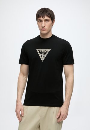 Homme portant un t-shirt noir à manches courtes avec un logo triangulaire beige Guess centré, associé à un pantalon beige clair, debout devant un fond uni.
