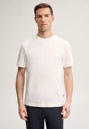 Homme aux cheveux bruns courts se tient face à l'avant, portant un t-shirt blanc uni à manches courtes et un pantalon noir sur un fond neutre.