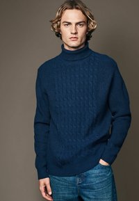 Dunkelblauer Kabelstrick-Pullover mit Rollkragen, gerippten Bündchen und Saum, getragen mit blauen Jeans, vor einem neutralen Hintergrund.