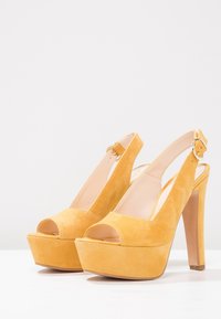 Žluté semišové platformové podpatky s otevřenou špičkou, designem na opasek, vysokým stiletto podpatkem a akcentem s přezkou. Hladká textura a zaoblený tvar.