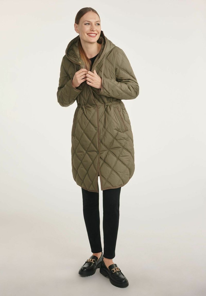 Olive-zielona pikowana kurtka puchowa z kapturem, z paskiem w talii, kieszeniami z przodu i brązowymi wykończeniami. Połączona z czarnymi spodniami i mokasynami.