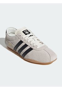 Gray suede sneaker with black stripes, beige tongue, and white laces. Features a gum sole and gold logo on the side.