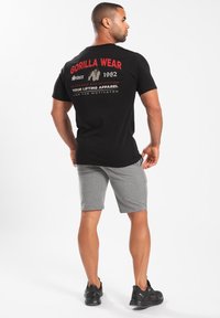 Zwarte T-shirt met korte mouwen en rode tekst en afbeelding op de rug, gecombineerd met grijze shorts. Model draagt zwarte sportschoenen.