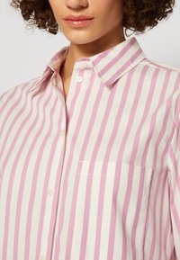 Femme portant une chemise blanche à rayures verticales roses, avec une poche poitrine et un col.