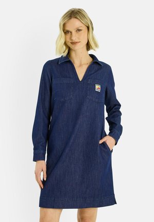 Mujer rubia vestida con un vestido largo de manga larga de mezclilla azul oscuro con bolsillos delanteros y mocasines negros con calcetines blancos, de pie frente a un fondo liso.