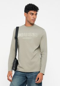 Cămașă gri deschis cu mânecă lungă, cu logo-ul embossat "GUESS" pe piept, asortată cu blugi din denim albastri. Modelul ține o geantă neagră tip crossbody.