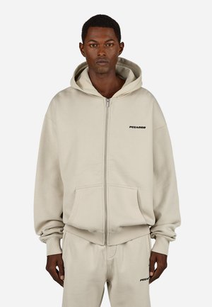 Homme portant un hoodie beige zippé et un pantalon de survêtement assorti avec de petits logos "PEGADOR" sur la poitrine et la cuisse, se tenant contre un fond blanc.