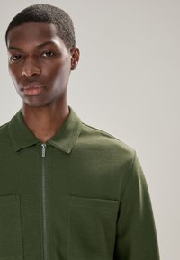 Chaqueta verde con cremallera y acabado texturizado, con cuello con solapa y dos bolsillos delanteros. Diseño sin costuras con detalles de construcción sutiles.