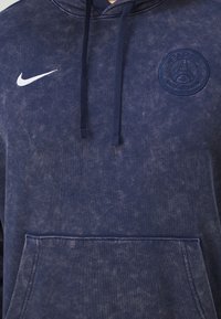 Granatowa bluza z kapturem wykonana z miękkiej tkaniny, z kangurzą kieszenią, sznurkami do regulacji i haftowanym logo Paryża. Biała sygnatura Nike na klatce piersiowej.