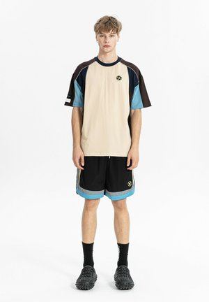 Joven de pie mirando hacia adelante, lleva una camisa de manga corta beige, negra y azul y pantalones cortos negros a juego con rayas azules y grises.