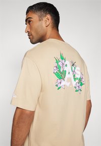 Camiseta de algodón beige con un gráfico floral colorido en la parte posterior, con hojas verdes y flores rosas, mangas cortas y cuello redondo.