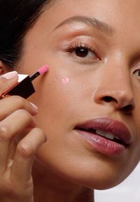 Du blush liquide dans une teinte rose pâle appliqué avec un petit applicateur sur la joue d'un mannequin. Les lèvres du mannequin sont d'un rose naturel ; l'arrière-plan est neutre.