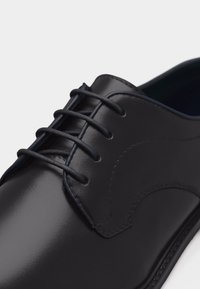 Κοντινό πλάνο ενός μαύρου γυαλισμένου δερμάτινου παπουτσιού, εστιασμένο στα κορδόνια και στη λεπτομέρεια ραφής στο πλάι.