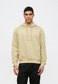 Felpa beige con cordino, polsi e orlo a costine. Realizzata in materiale morbido, presenta una vestibilità rilassata e un design semplice. Abbinata a pantaloni neri.