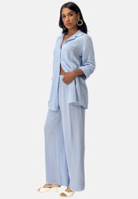 Lichtblauwe, gestructureerde, oversized blouse met opgerolde mouwen gecombineerd met bijpassende losse broek. Open voorkant, gedragen met witte hakken sandaaltjes.
