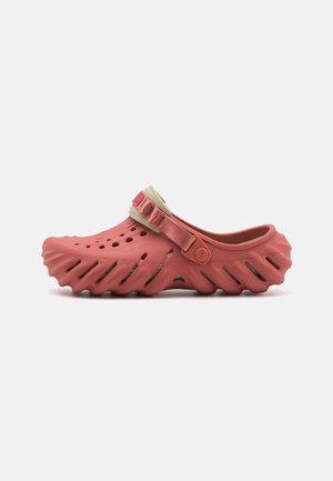 Red rubber clog with a textured, wavy sole, perforations for breathability, and a beige adjustable strap for secure fit.